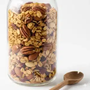 Gros plan d’un bocal en verre transparent contenant du granola maison sans sucre, avec des flocons d’avoine dorés, des amandes, des noix de pécan et des cranberries. Fond blanc épuré avec une cuillère en bois à côté, soulignant la texture croustillante et les couleurs naturelles pour une fiche recette claire."