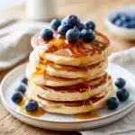 Pancakes sans sucre moelleux garnis de myrtilles fraîches et d'un filet de miel, servis sur une assiette blanche